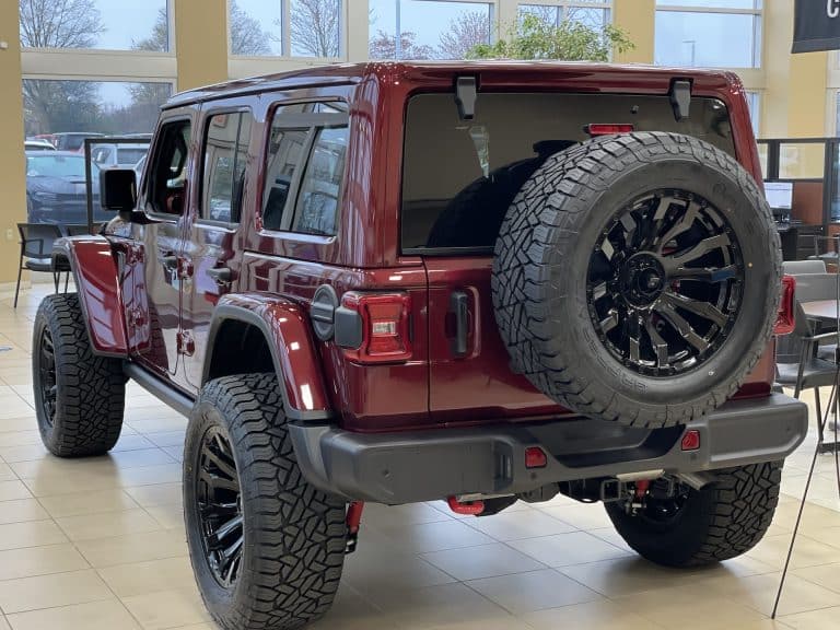 A maroon Jeep with black rims is parked indoors, showcasing its spare tire mounted on the back. The vehicle is positioned at an angle, highlighting its rear and side, inside a showroom with large windows in the background.