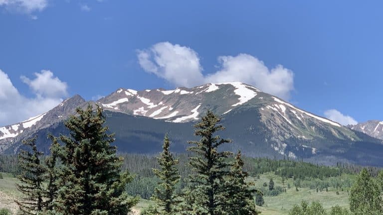 Snow-capped mountain under a clear blue sky, with green pine trees and lush meadows in the foreground. Ideal for mountain travel getaways, white fluffy clouds drift above the peak, promising adventure and serenity.