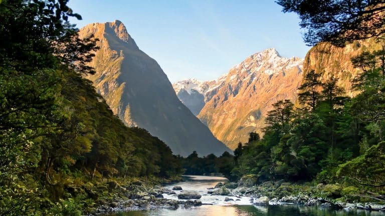 A serene river flows through a lush forest, flanked by towering, rugged mountains with snow-capped peaks under a clear blue sky. The scene is illuminated by warm sunlight, highlighting the natural beauty of the landscape.