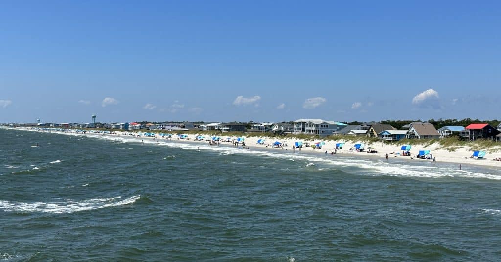 A scenic beach view on a sunny day, with rows of beach umbrellas and chairs along the shoreline. People are scattered across the sand, enjoying the seaside. In the background, houses are visible against a clear blue sky with a few clouds. Waves crash gently on the shore.