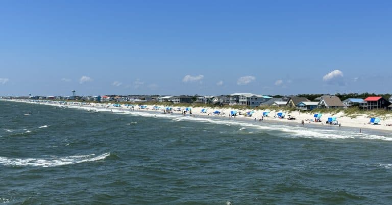 A scenic beach view on a sunny day, with rows of beach umbrellas and chairs along the shoreline. People are scattered across the sand, enjoying the seaside. In the background, houses are visible against a clear blue sky with a few clouds. Waves crash gently on the shore.