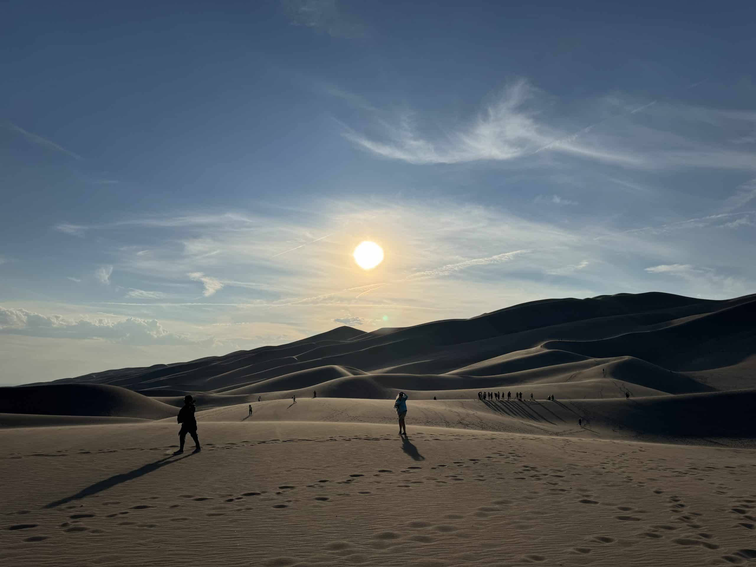 A vast desert landscape at sunset with sand dunes and scattered footprints. Silhouetted figures are visible walking on the sand, casting long shadows. The sky is mostly clear with a few clouds, and the sun is low on the horizon.