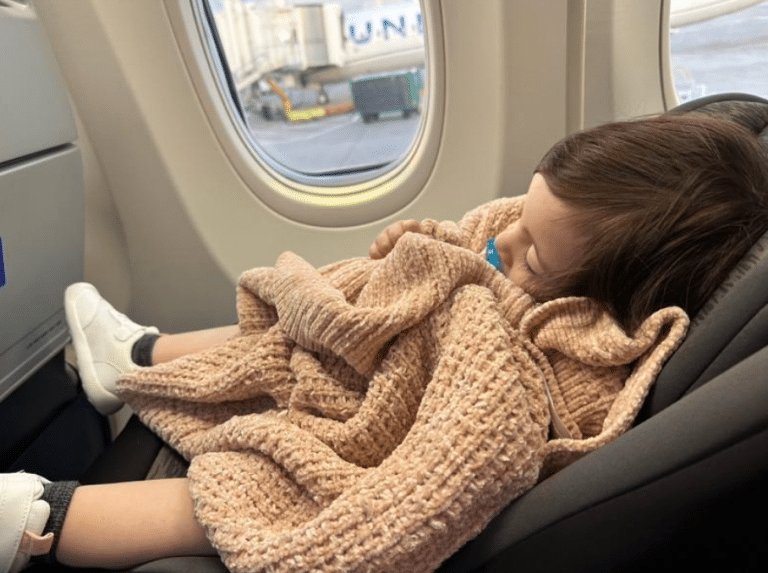 A young child sleeps in a car seat on an airplane, wrapped in a thick, pink knitted blanket. The child's head leans to the side, with a pacifier in their mouth. The airplane window next to the child shows the view of airport ground equipment outside.