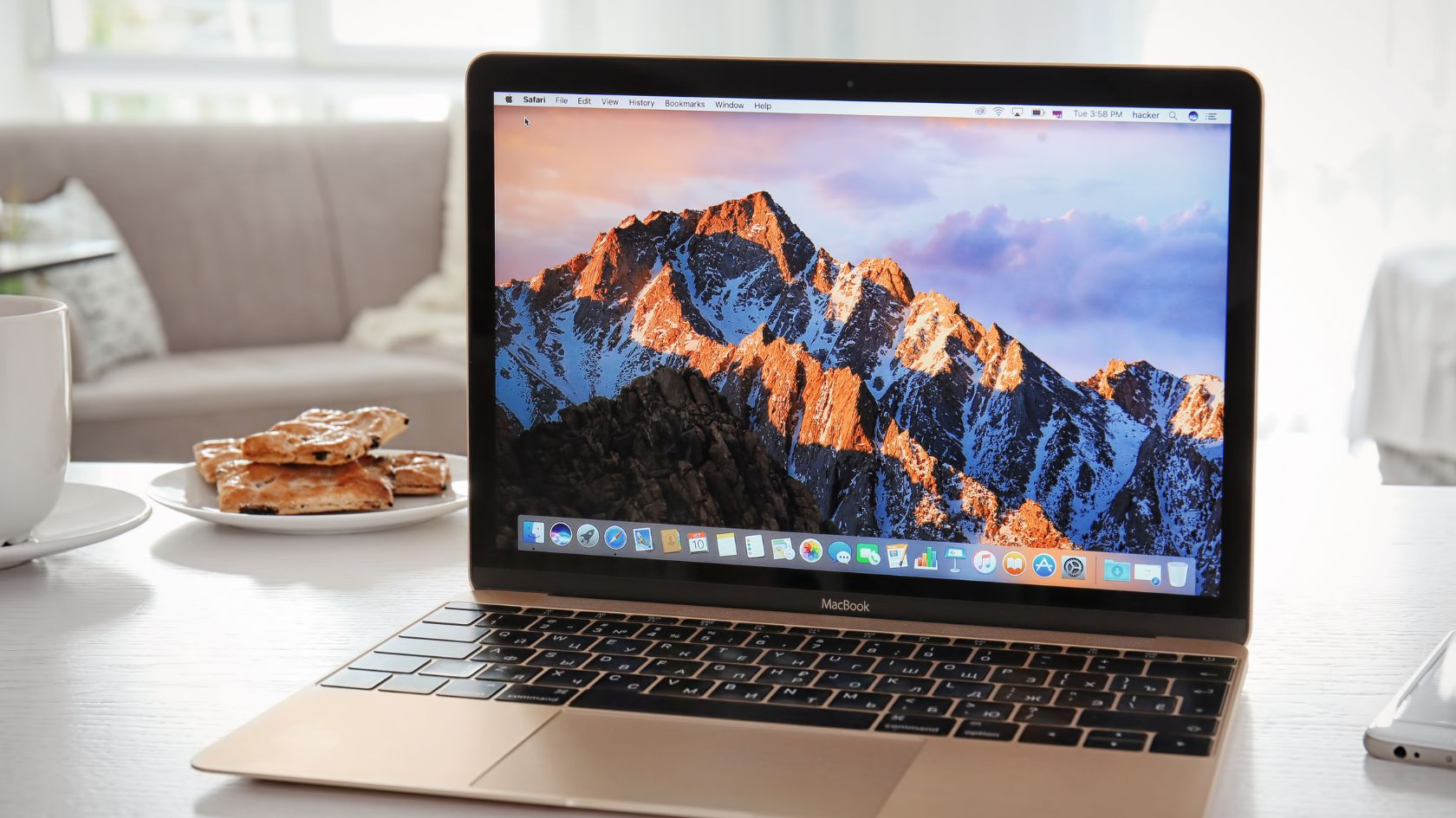 A MacBook with a mountain landscape wallpaper is open on a white table. The screen displays multiple application icons. There's a plate of pastries and a white cup on the table. In the background, a light-colored couch and a window with sheer curtains are visible.