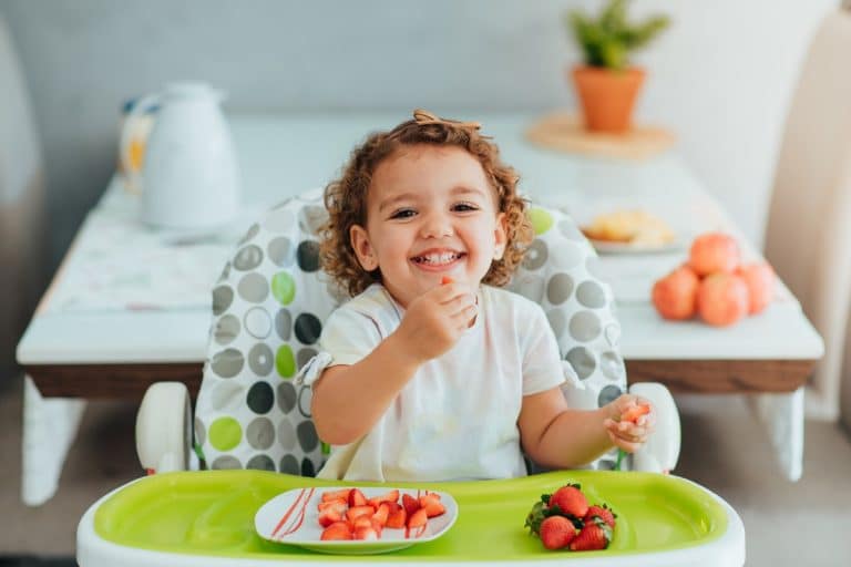 A joyful toddler with curly hair sits in a high chair, holding a strawberry in one hand and smiling. In front of the child is a plate with cut strawberries. The background features a table with a plant and oranges.