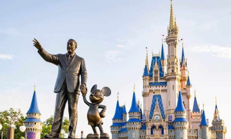 A bronze statue of a man holding hands with a mouse character stands in front of a castle with blue and gold spires at what appears to be a theme park. The clear sky adds to the picturesque scene.