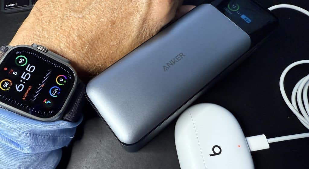 A wrist adorned with a smartwatch and its vibrant display reads 6:55. Beside it lies a silver Anker power bank and sleek white earbuds with a 'b' logo charging case, all essential gadgets for tech-savvy travel enthusiasts.