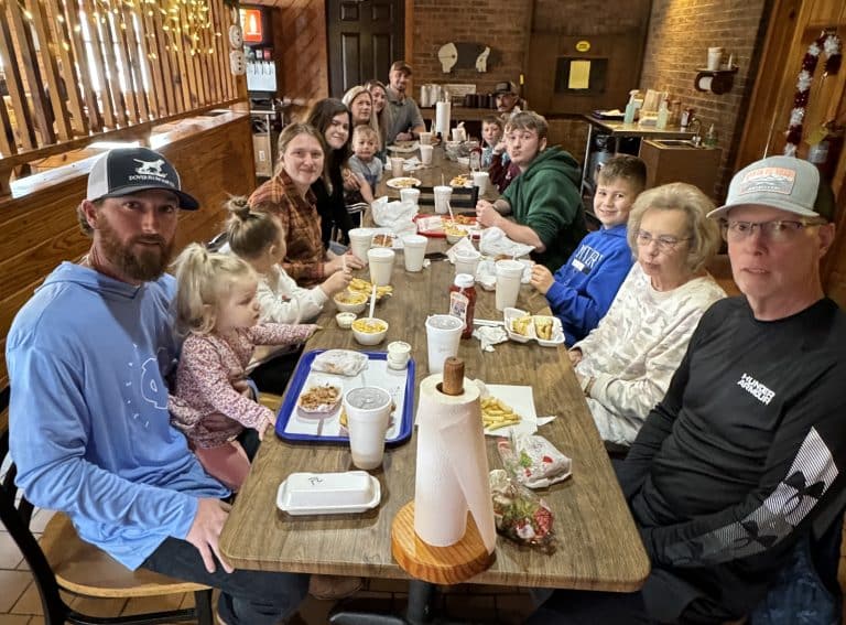 A large family, spanning multiple generations, is gathered around a long wooden table in a restaurant. They are enjoying a meal together with various dishes and drinks in front of them. The setting has a cozy, rustic ambiance with warm lighting.