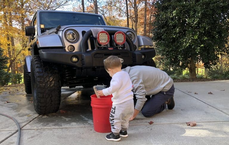 A toddler in a white sweater and gray sweatpants holds a brush next to a red bucket by an off-road vehicle equipped with top smart auto gadgets. An adult, partially visible, kneels beside the child amidst trees adorned with autumn leaves.