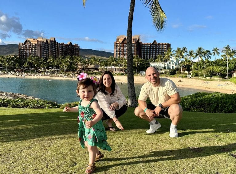 A cheerful family is enjoying a sunny day at the beach. A young child, wearing a floral dress, runs in the foreground. Behind, a woman and a man crouch on the grass, smiling. Palm trees and a resort are visible in the background.